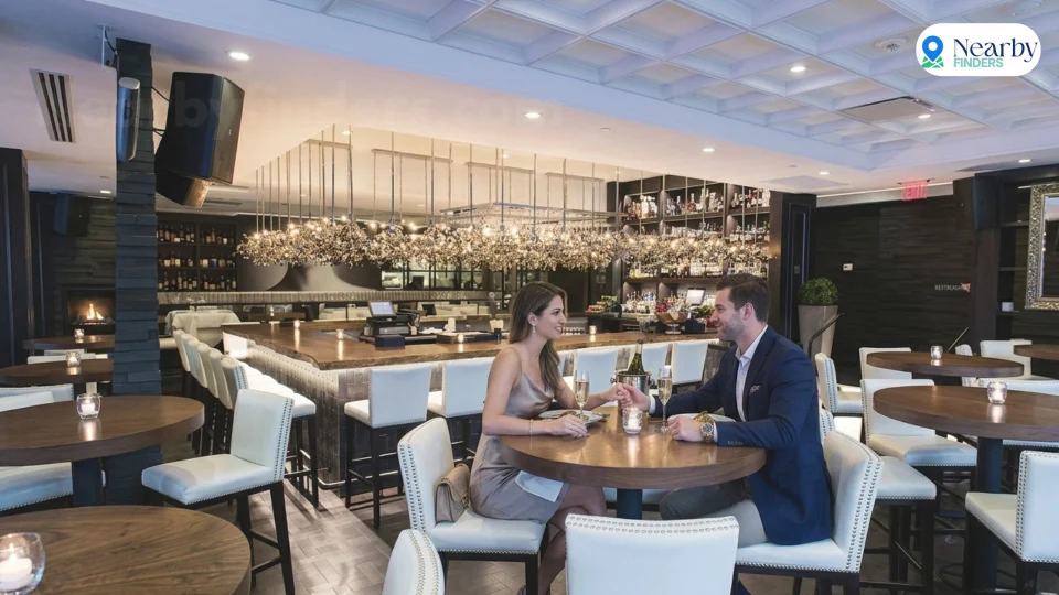 Romantic restaurants for couples in Houston: A glamorous couple holding hands at a round table near the bright, luxury white leather chairs and lit bar inside Steak 48 Houston